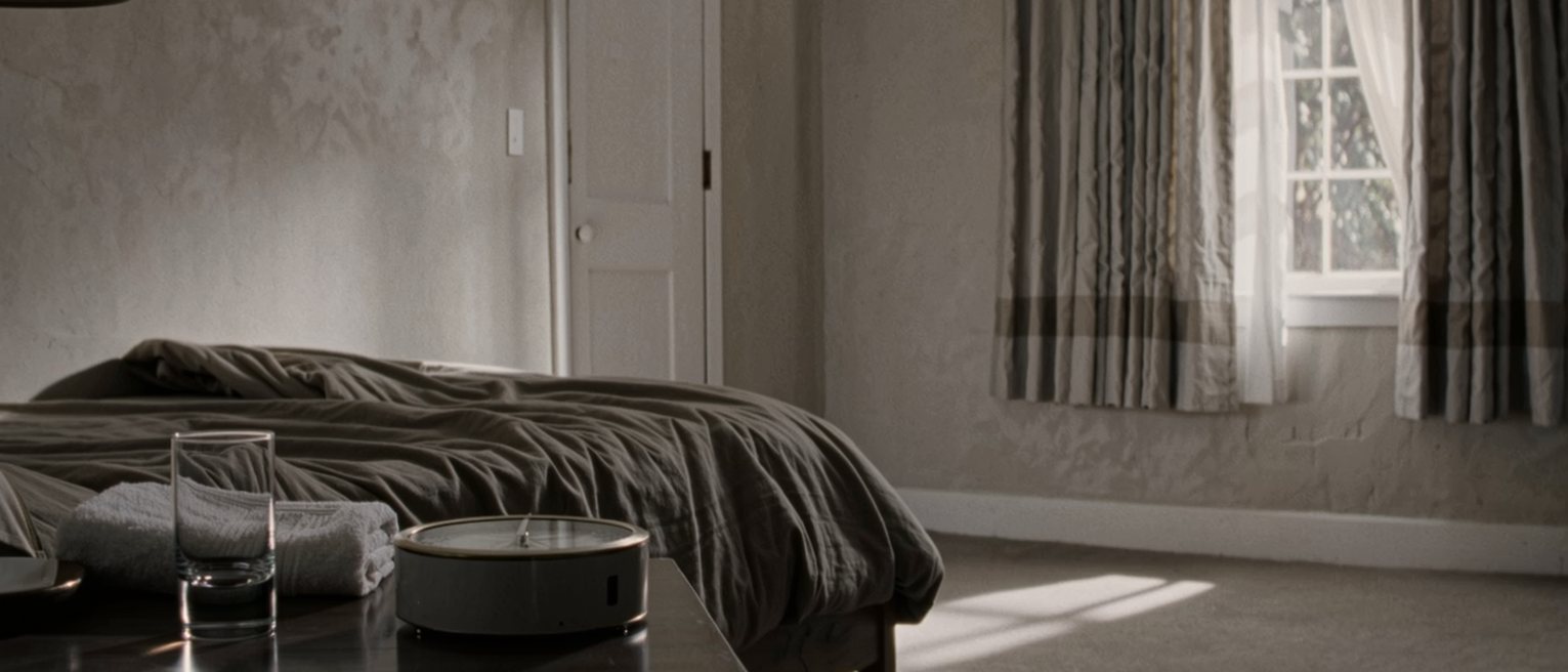 a bedroom seen close with a small clock unplugged and laid face down on the bedside table, a glass of water and a folded towel, the bed with its cover turned back close behind and the curtains drawn beyond