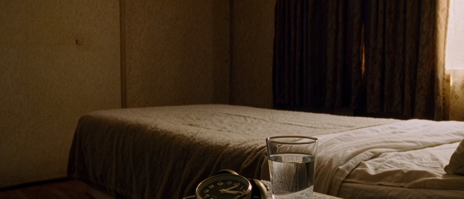 a bedroom seen close with a small clock unplugged and laid face down on the bedside table, a glass of water and a folded towel, the bed with its cover turned back close behind and the curtains drawn beyond