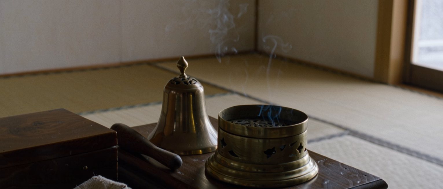 a tatami room seen close with a low table holding a brass incense burner with its smoke gone out, a small bell and its striker beside it, a folded cloth and a closed wooden box, the shoji soft beyond