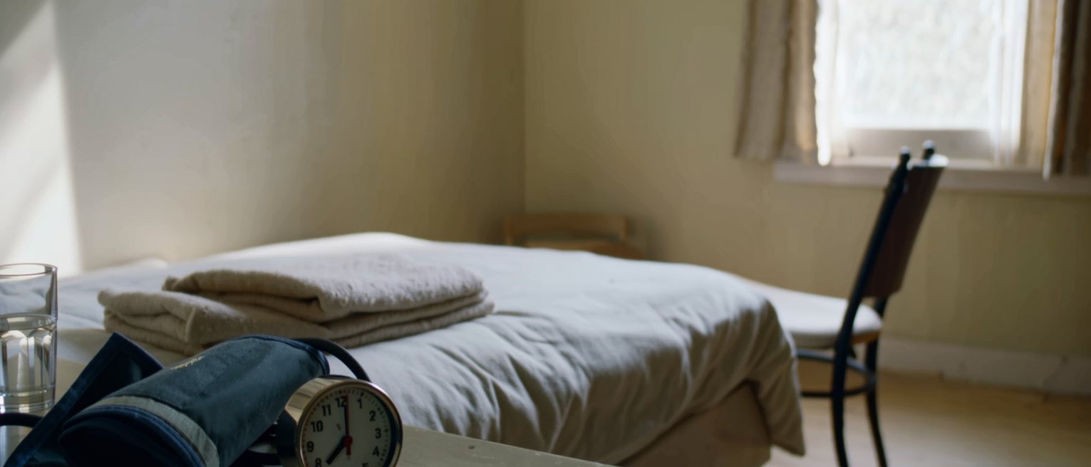 a bedside seen close with a blood pressure cuff coiled beside a small clock and a glass of water on the table, a folded blanket at the foot of the bed, a chair drawn up, the window soft beyond