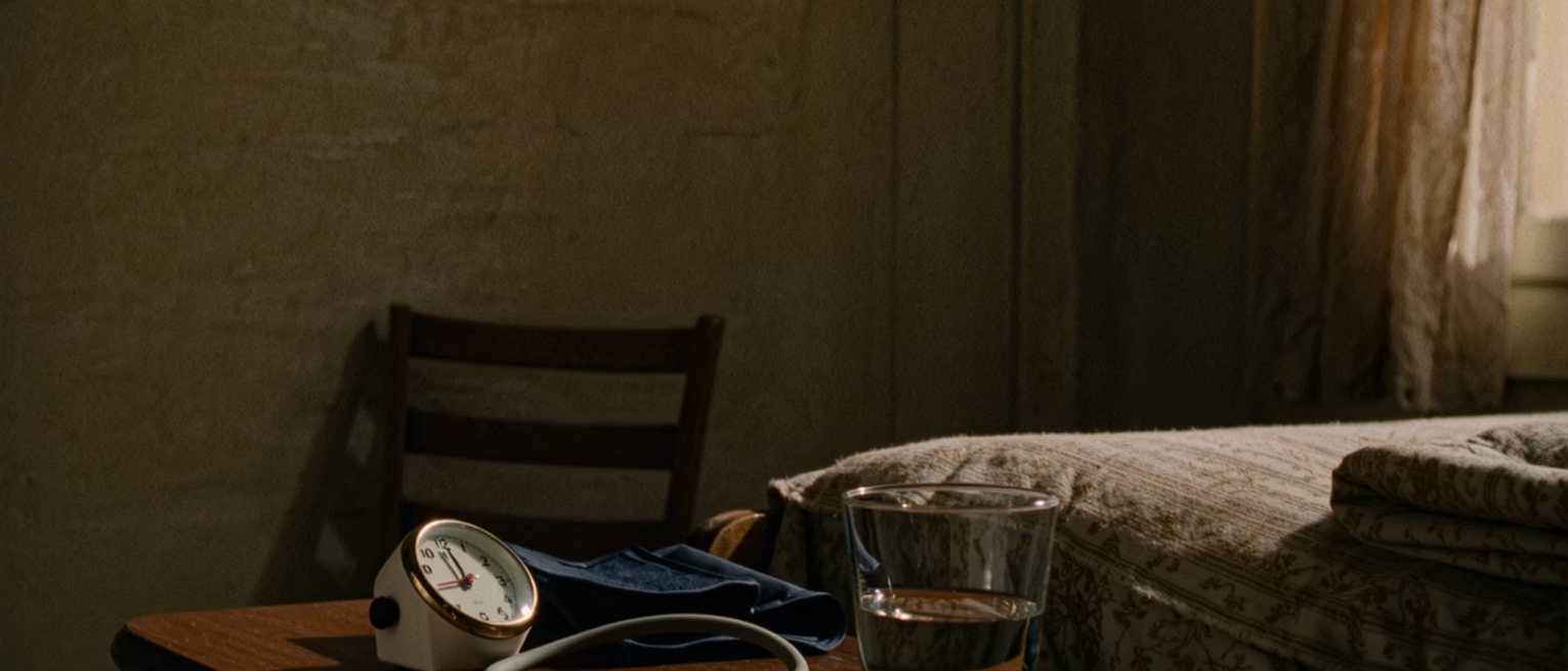 a bedside seen close with a blood pressure cuff coiled beside a small clock and a glass of water on the table, a folded blanket at the foot of the bed, a chair drawn up, the window soft beyond