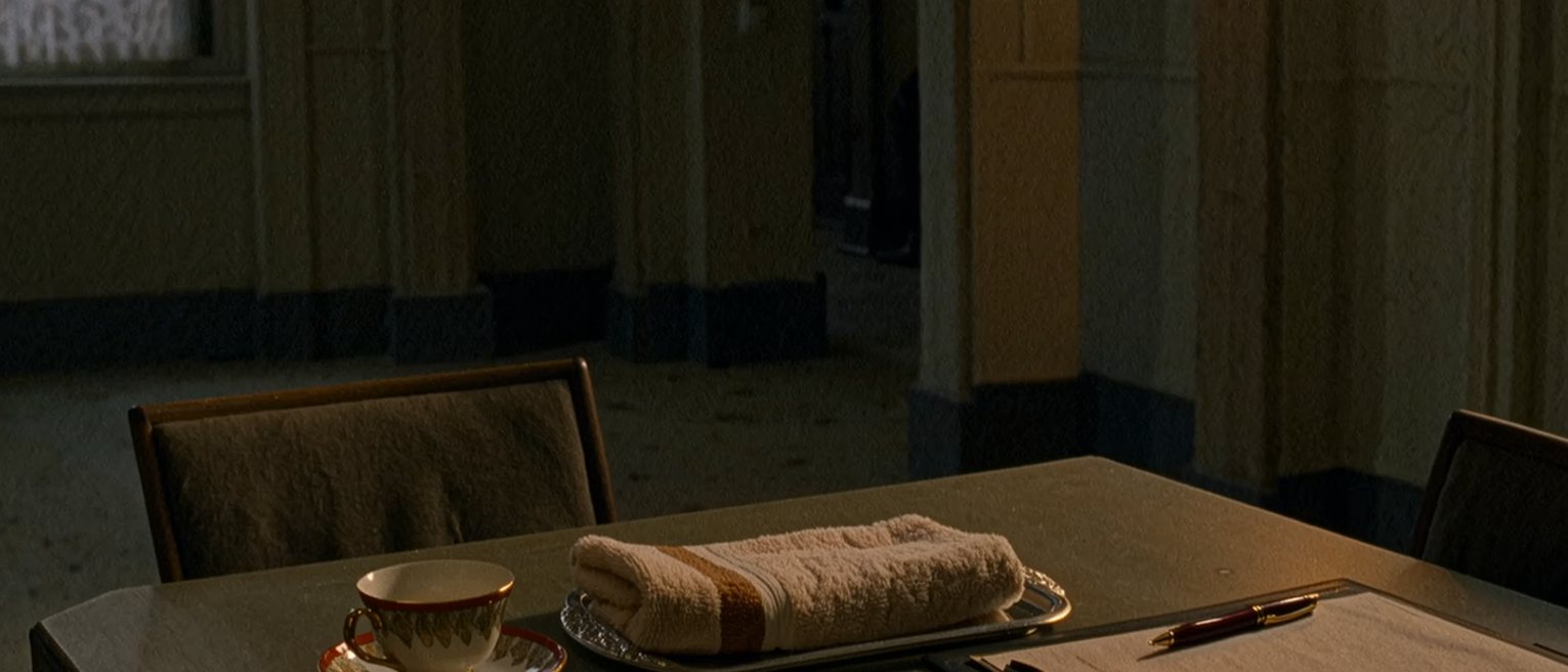 a table seen close with three objects set apart in a row: a second teacup, a folded warm towel on a small tray, a document folder with a pen across it, the chairs pushed in, the window soft beyond