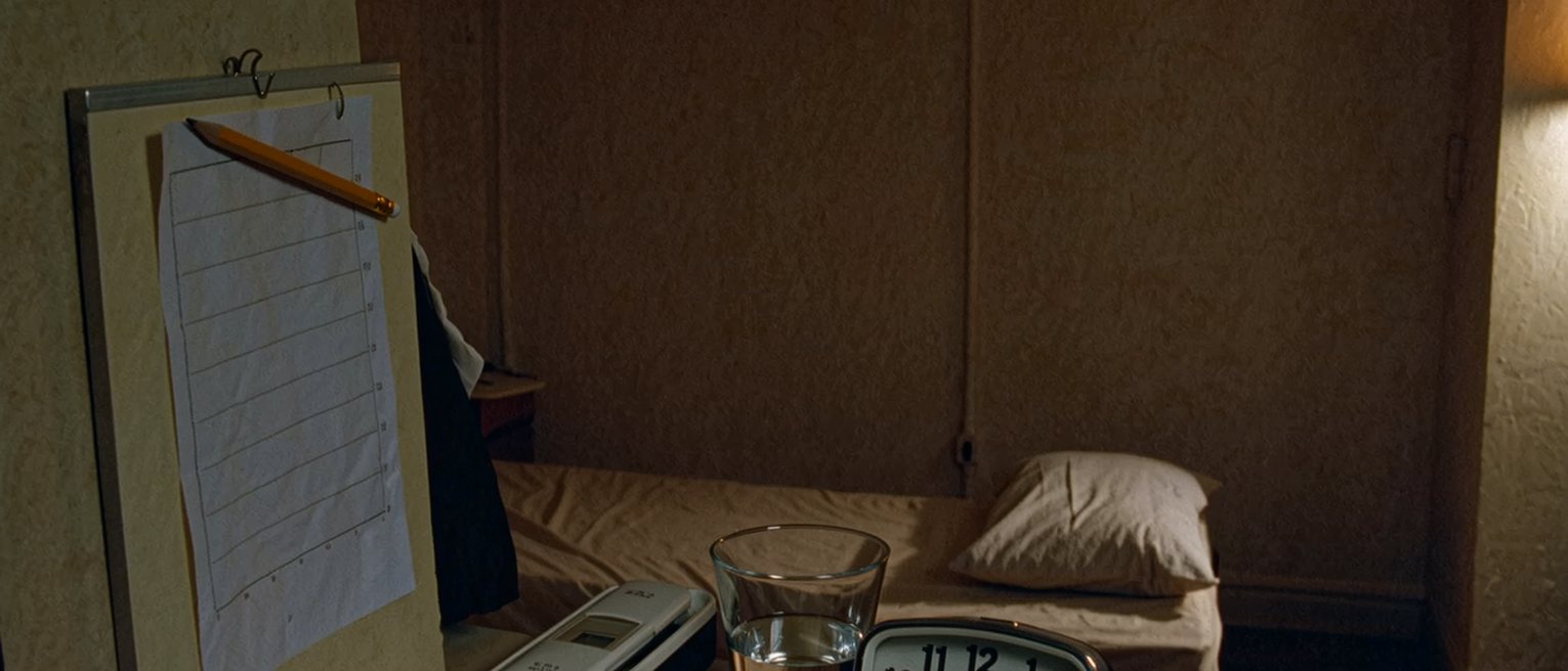 a bedside seen close with a chart clipped to a small board and a pencil tied to it, a thermometer in its case, a glass of water and a clock, the bed turned back behind