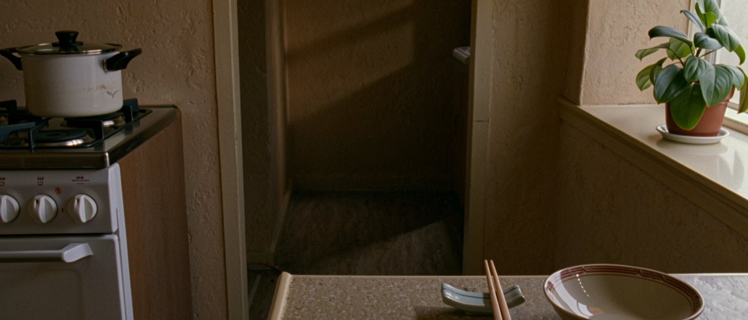 a small apartment kitchen seen close with one place laid at the counter, one bowl and one pair of chopsticks on a rest, a single pot on the stove, a plant on the sill, the window soft beyond