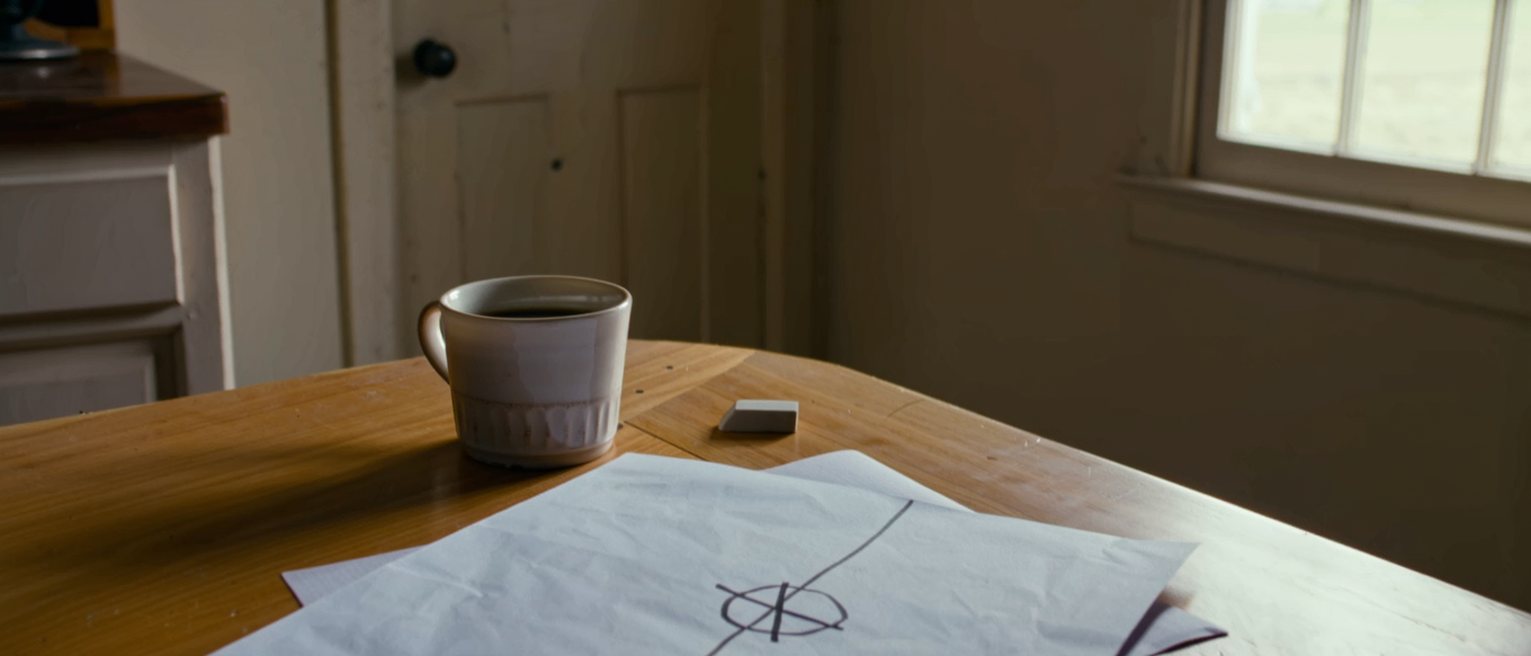 a kitchen table seen close with a sheet of paper on which a straight line and a circle are drawn in pencil, one crossed out, a cup of coffee and an eraser, the dresser close behind and the window soft beyond
