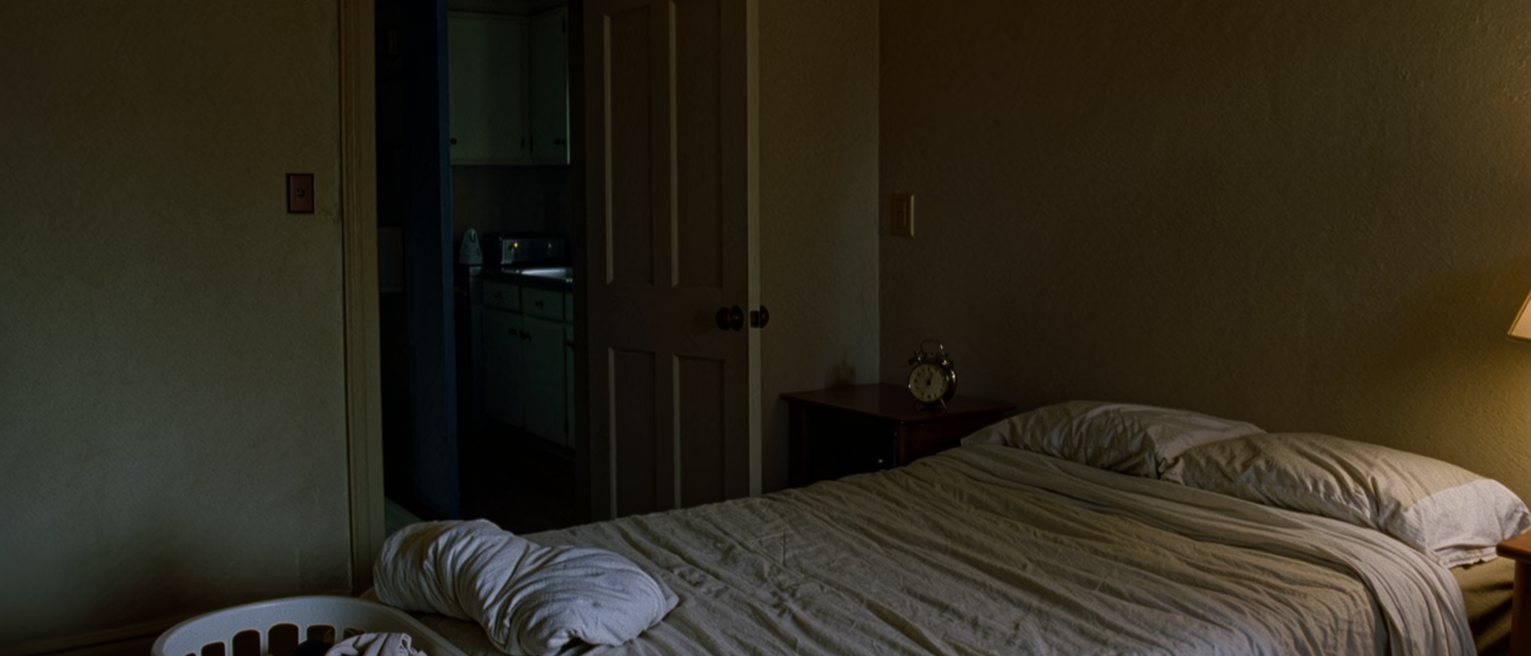 a bedroom seen close with one side of the bed slept in and the other side still made, an alarm clock on the near table, a full laundry basket at the foot of the bed, the door ajar onto the kitchen beyond