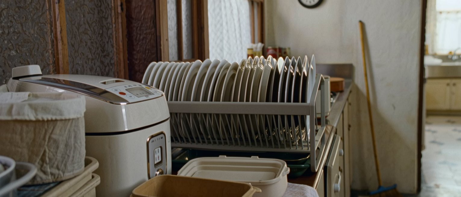 a kitchen seen close with a rack of washed dishes, a rice cooker with its timer set, an empty lunch box laid open on the counter beside its cloth, a wall clock, the window soft beyond