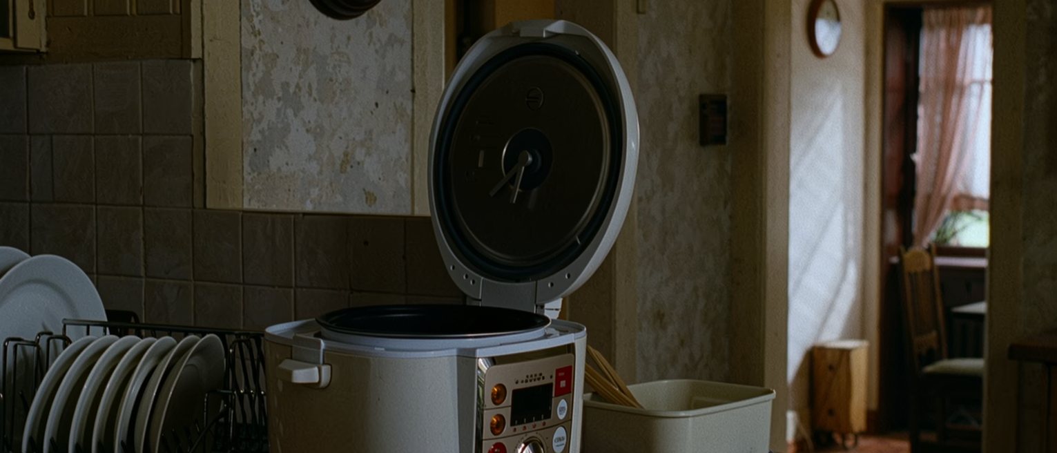 a kitchen seen close with a rack of washed dishes, a rice cooker with its timer set, an empty lunch box laid open on the counter beside its cloth, a wall clock, the window soft beyond