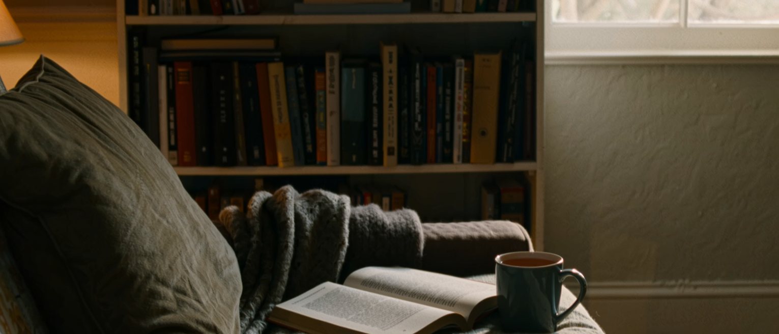 a living room seen close with a soft blanket folded over the arm of the sofa, a cushion still dented, a mug of tea and a paperback, the bookshelf close behind and the window soft beyond