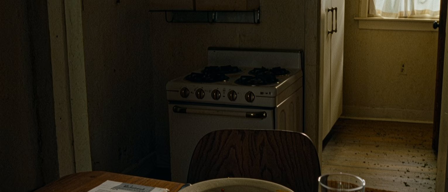a kitchen table seen close with a booking confirmation printed and folded beside a plate with the remains of dinner, a fork laid down and a glass of water, the stove close behind and the window soft beyond