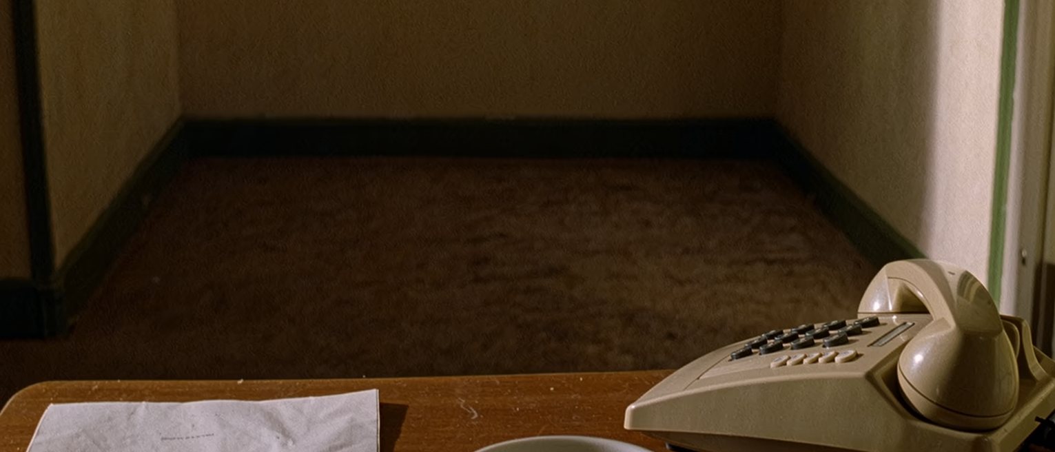 a table seen close with a folded printed sheet squared beside a pen that has not been used, a cup of tea gone cold, a telephone resting on its cradle, the window soft beyond