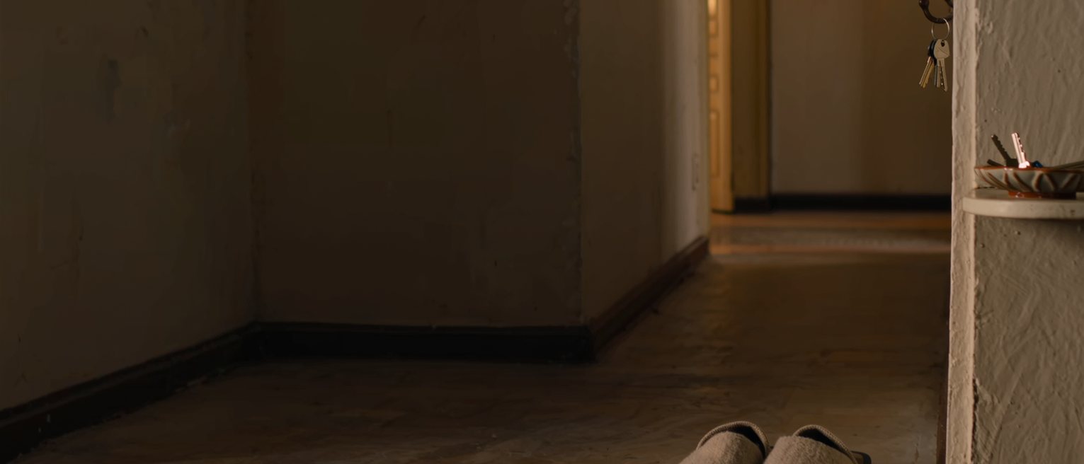 a hall seen close with a pair of slippers set out square by the step, an empty hook above, a small dish of keys on the shelf, the wall soft beyond
