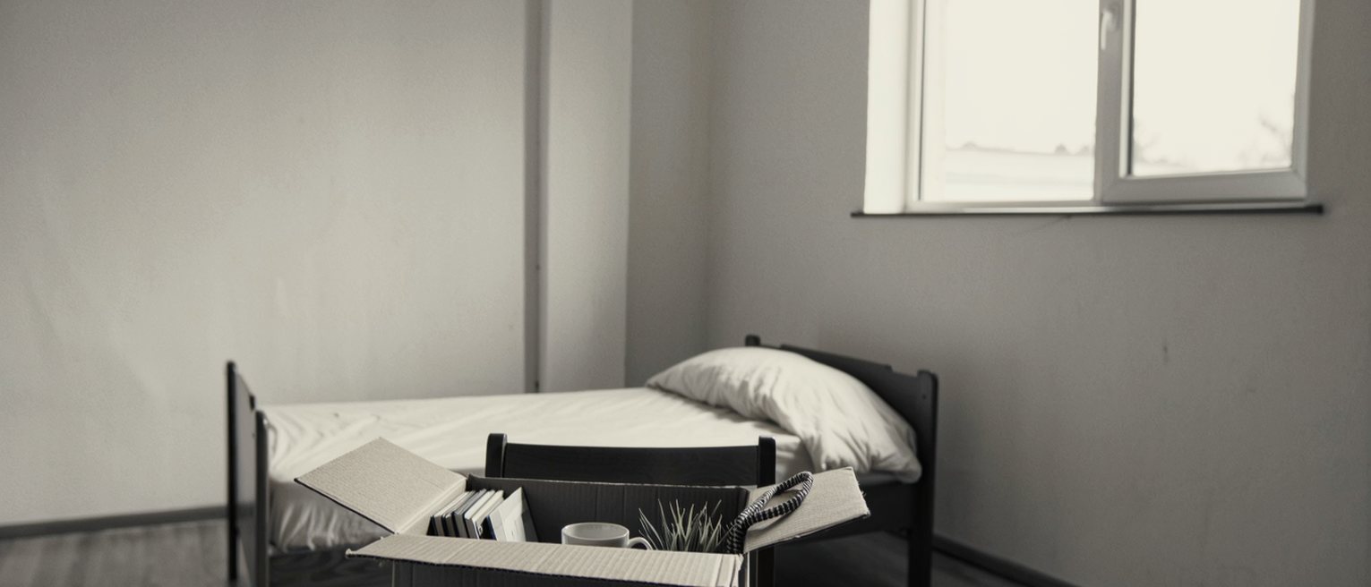 a bedroom seen close with a cardboard box set on the chair holding a desk tidy, a mug and a small potted plant, a lanyard coiled on top, the bed made behind, the window soft beyond