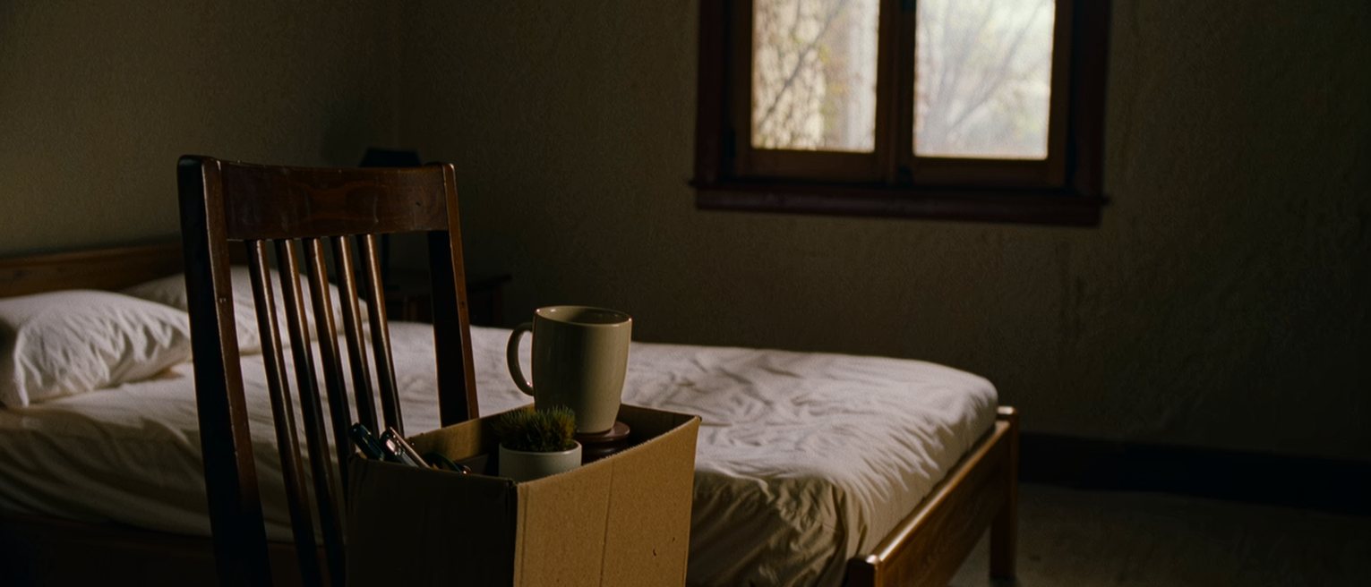 a bedroom seen close with a cardboard box set on the chair holding a desk tidy, a mug and a small potted plant, a lanyard coiled on top, the bed made behind, the window soft beyond