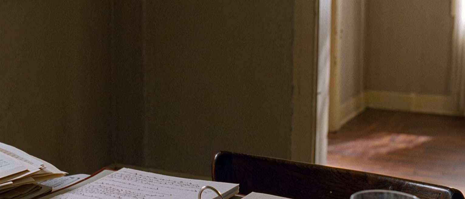 a table seen close with a ring binder open at a run of dated notes in pen, a stack of printed sheets in a clear sleeve beside it, a cup gone cold, the chair pushed in, the window soft beyond