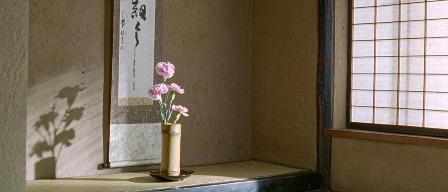 a tokonoma seen close with a single stem of fringed pink in a bamboo vase, a hanging scroll of brush calligraphy behind it, a small dish of water, the alcove post close behind and the shoji soft beyond