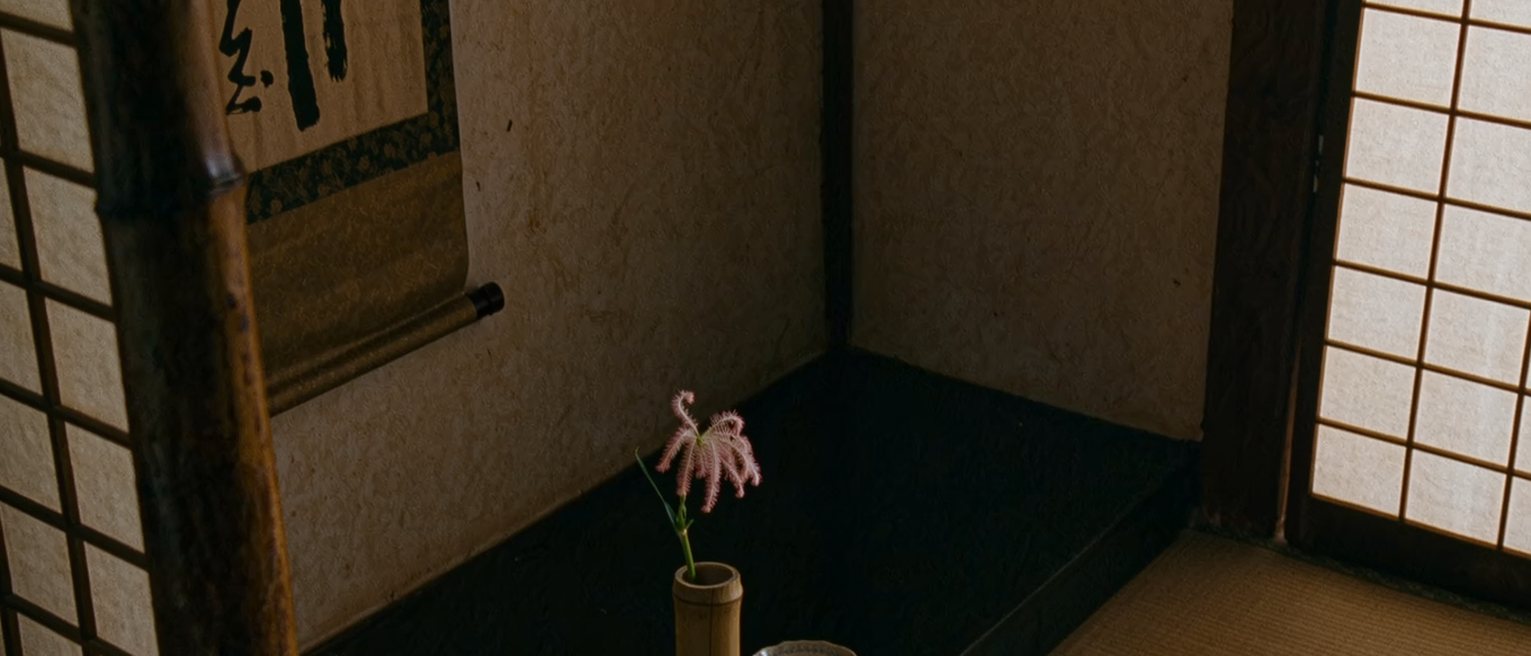 a tokonoma seen close with a single stem of fringed pink in a bamboo vase, a hanging scroll of brush calligraphy behind it, a small dish of water, the alcove post close behind and the shoji soft beyond