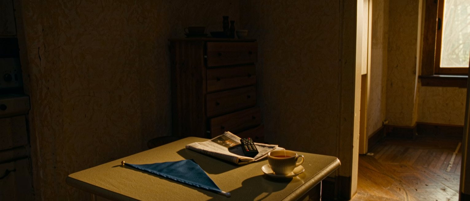 a kitchen table seen close with a small blue pennant laid flat, a folded newspaper, a television remote and a cup of tea gone cold, the dresser close behind and the window soft beyond