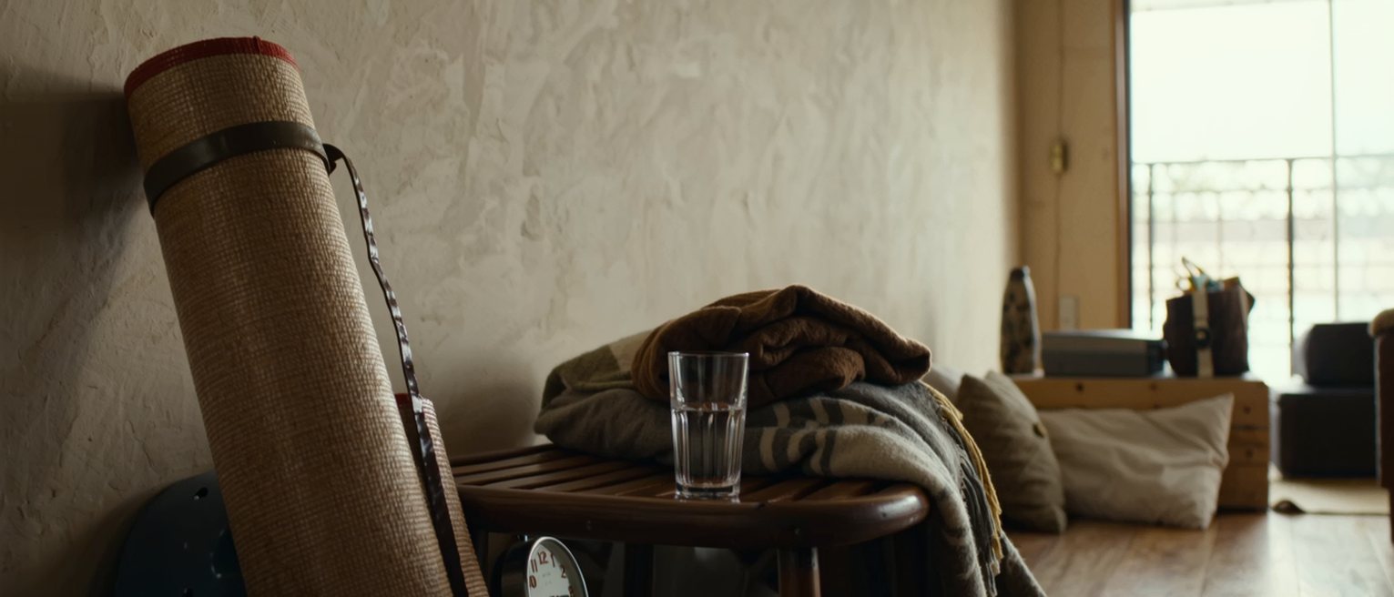 a sitting room seen close with a rolled mat standing against the wall beside a folded blanket on the low stool, a glass of water, a clock, the room soft beyond
