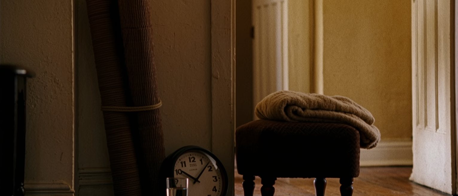 a sitting room seen close with a rolled mat standing against the wall beside a folded blanket on the low stool, a glass of water, a clock, the room soft beyond