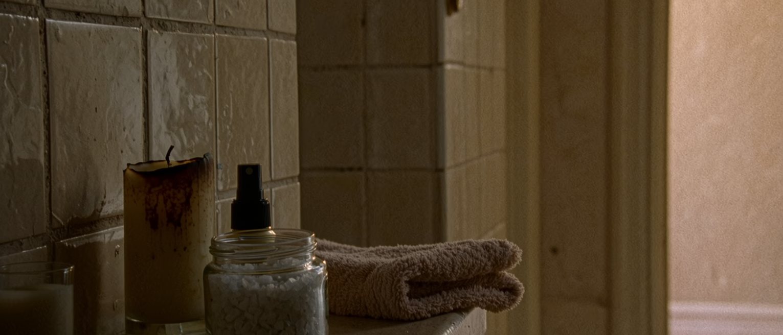 a bathroom shelf seen close with a candle burnt down in its glass, a jar of bath salts with the lid off, a small spray bottle and a folded flannel, the tiled wall soft beyond