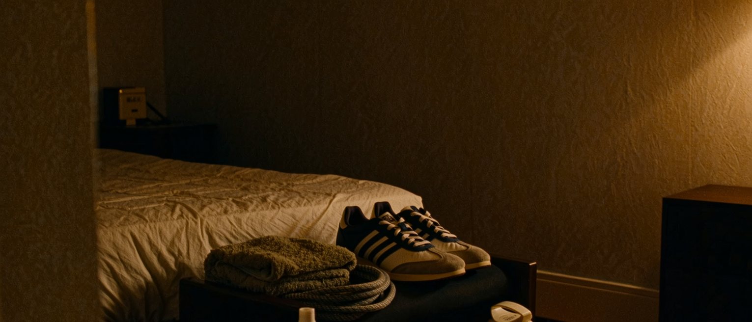 a bedroom seen close with a pair of trainers set out on the low stool beside a coiled skipping rope and a folded towel, a water bottle, the bed made behind, the window soft beyond
