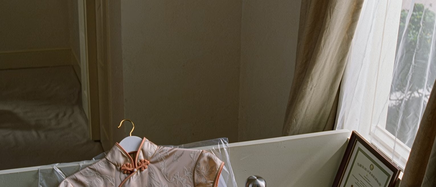 a dressing room seen close with a cheongsam on a hanger under a plastic cover, a prize certificate framed and one frame left empty beside it, a thermos and a pair of heels, the curtain soft behind