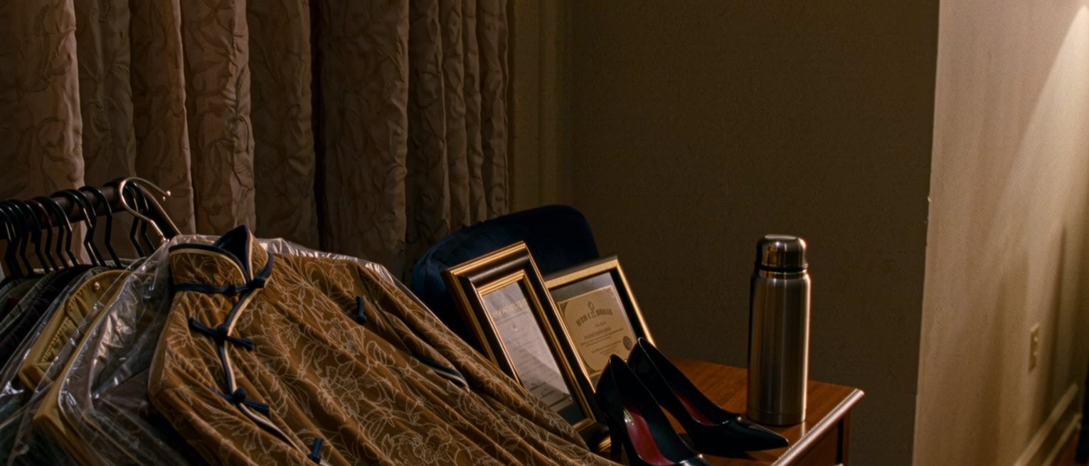 a dressing room seen close with a cheongsam on a hanger under a plastic cover, a prize certificate framed and one frame left empty beside it, a thermos and a pair of heels, the curtain soft behind