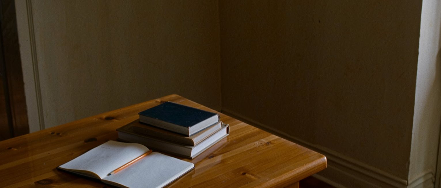a kitchen table with a notebook open at a blank page and a pencil laid in the fold, a stack of closed books beside it