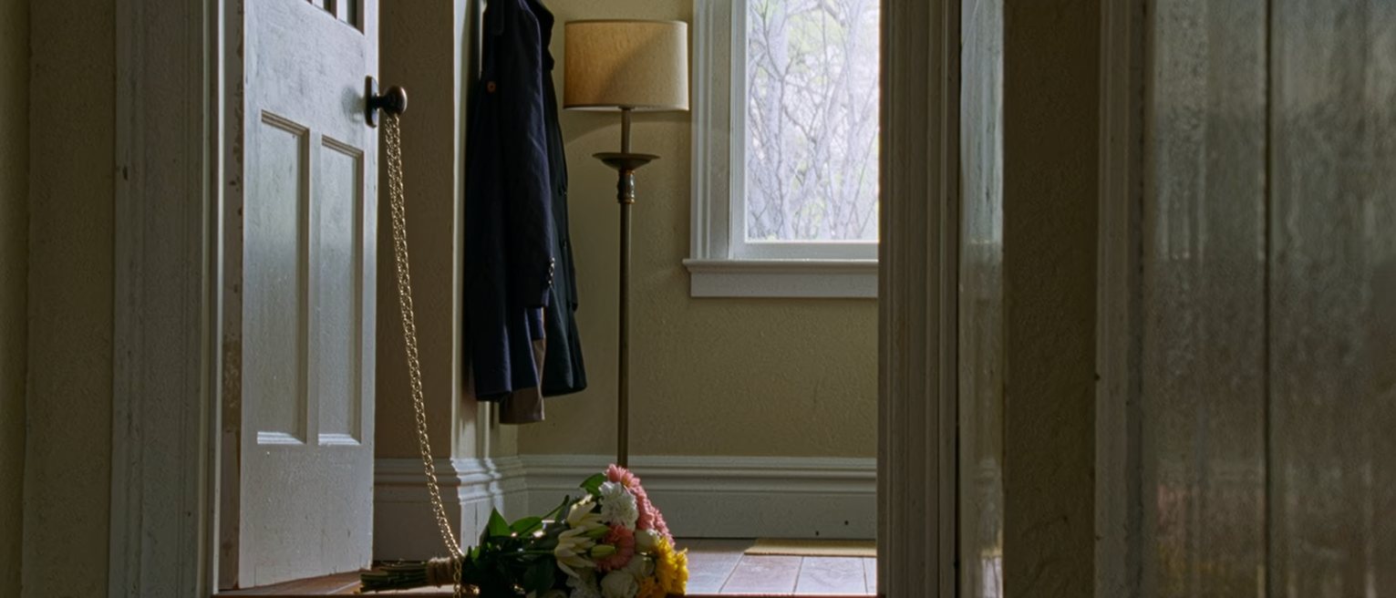 a front door with the chain on and a bunch of flowers left outside on the step, seen through the gap from inside, coats on the hooks and the hall lamp close beside the door