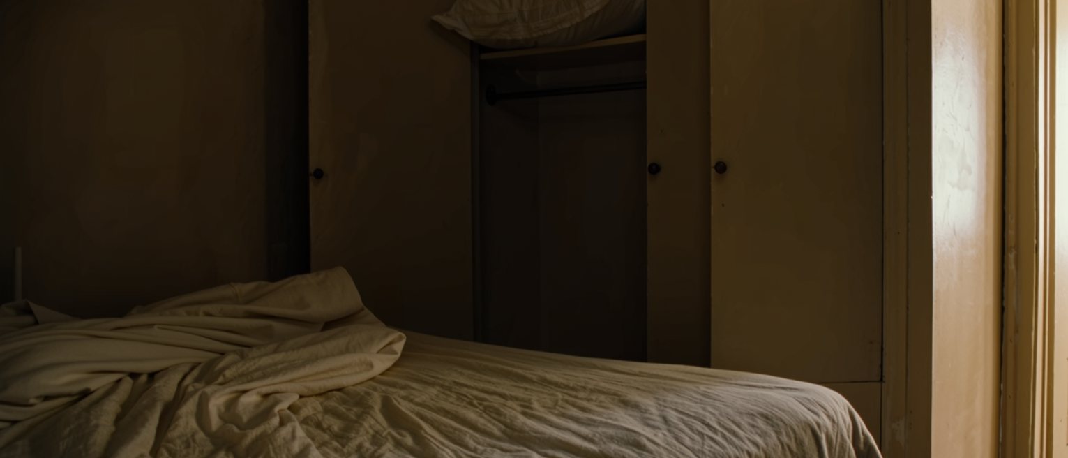 a bedroom with the covers turned down on one side only, the second pillow put away on the wardrobe shelf, a lamp and a book on the near bedside table