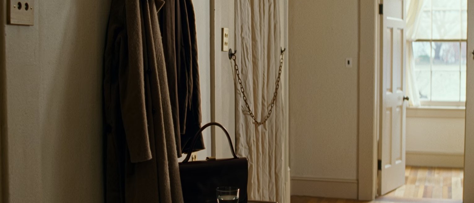 a hall seen close with a coat back on its hook, a clutch bag on the console beside a phone face down and a single glass of water, the door with its chain across soft behind