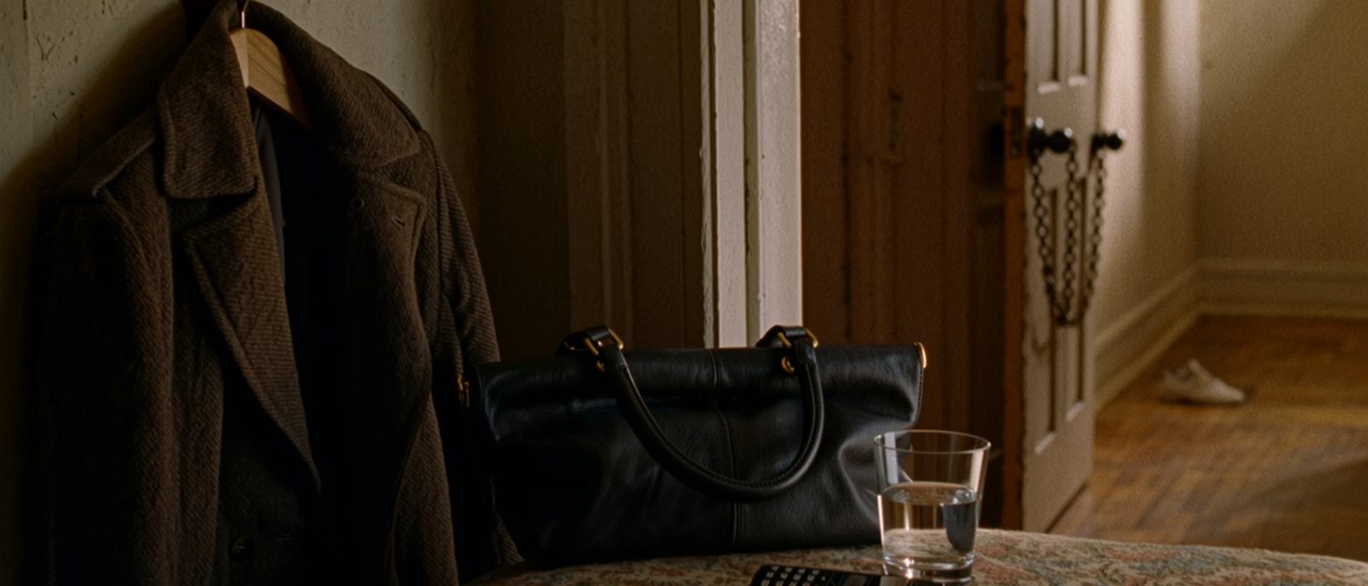 a hall seen close with a coat back on its hook, a clutch bag on the console beside a phone face down and a single glass of water, the door with its chain across soft behind