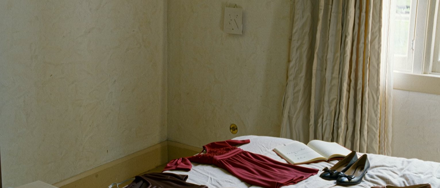 a bedroom seen close with two dresses laid on the bed and one chosen, a pair of flat shoes beside a pair of heels, a phone charging and a notebook open at a written line, the curtain close behind