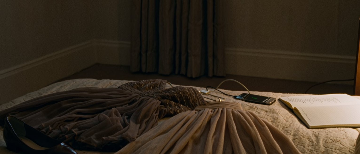 a bedroom seen close with two dresses laid on the bed and one chosen, a pair of flat shoes beside a pair of heels, a phone charging and a notebook open at a written line, the curtain close behind