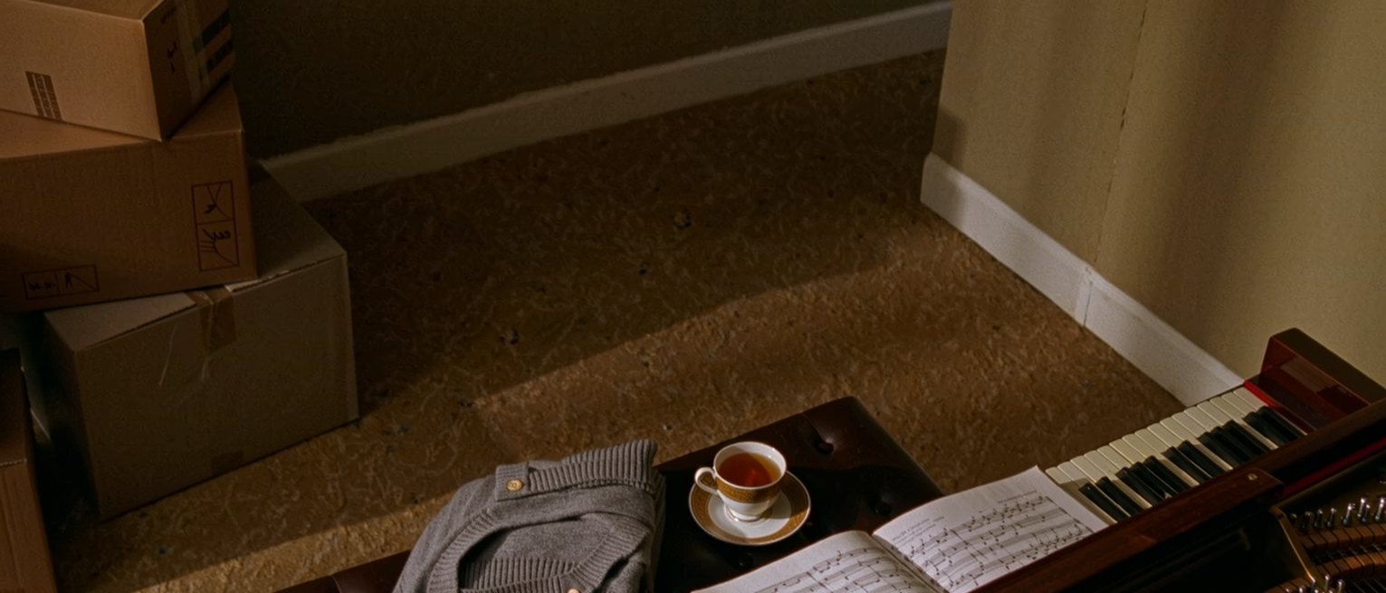 a sitting room seen close with a piano bench and a piano primer open on the stand, a cardigan folded on the bench and a cup of tea gone cold, the boxes from the move still stacked soft behind