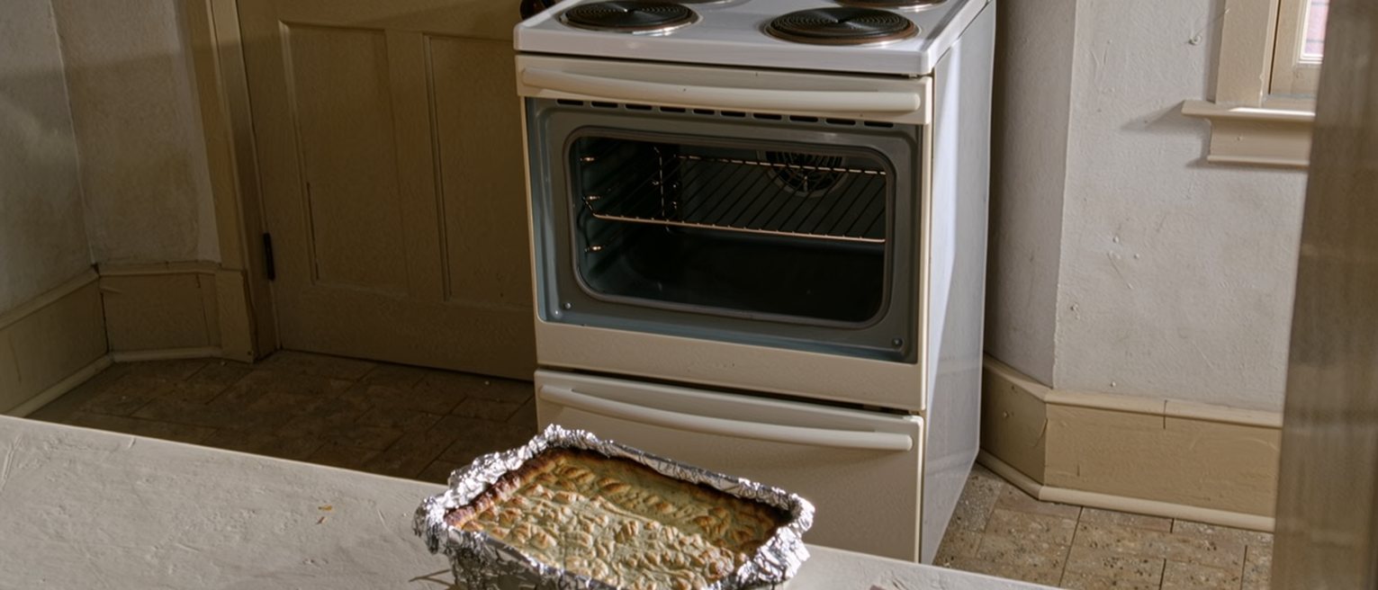 a kitchen counter where a casserole dish brought round from next door still sits covered in foil beside a folded note, untouched, the oven empty behind it
