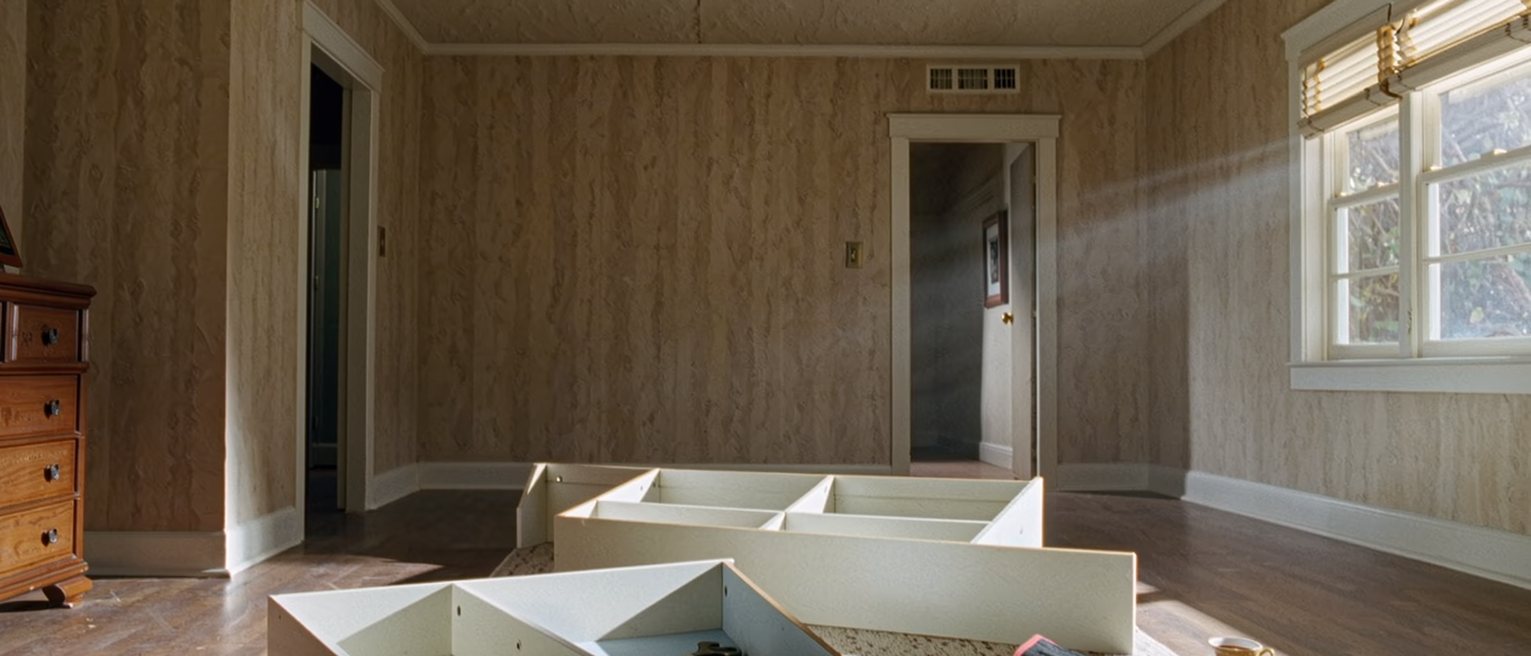 a sitting room where a bookshelf has been carried in and left unassembled, its parts laid out on the rug beside an open toolbag and a second cup of tea