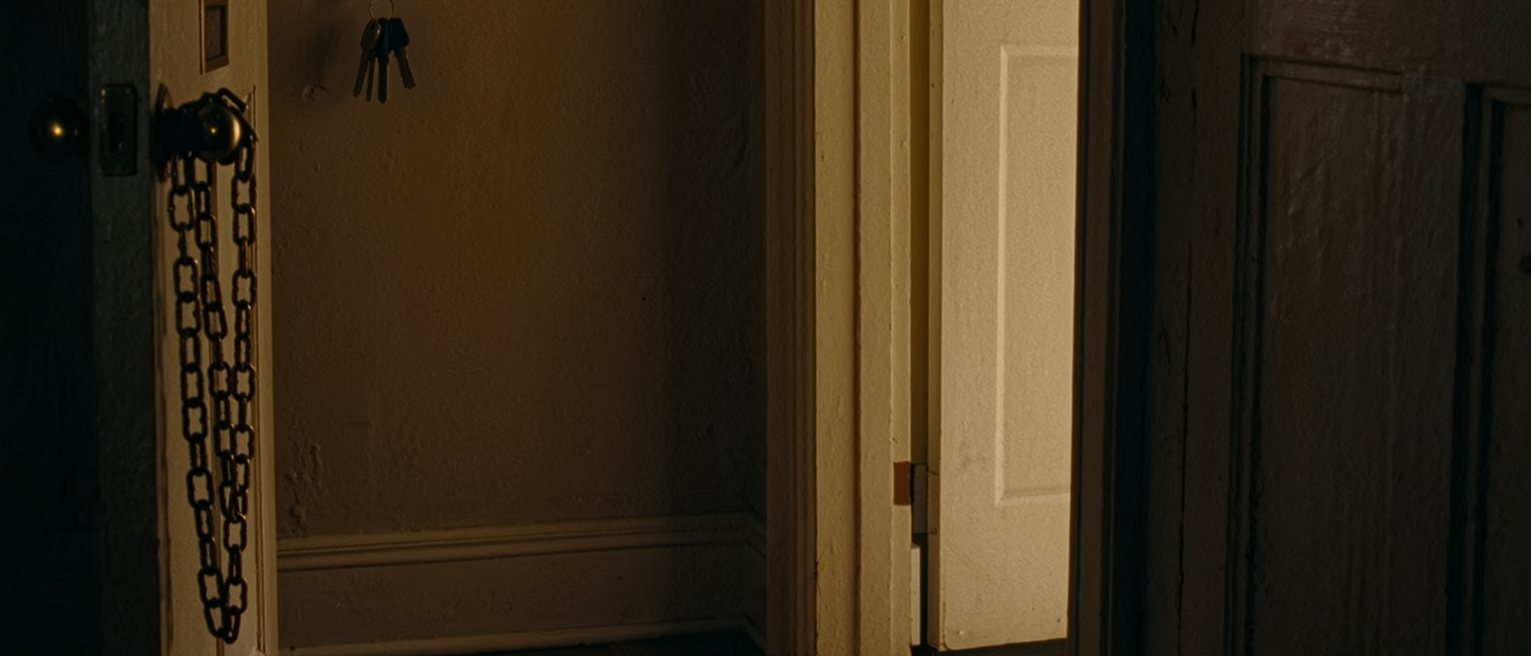 a hallway by the front door: a second set of keys hanging on a hook beside the first, a spare pair of slippers set out on the mat beneath, coats on the rack close behind and the door standing straight, its chain hanging loose