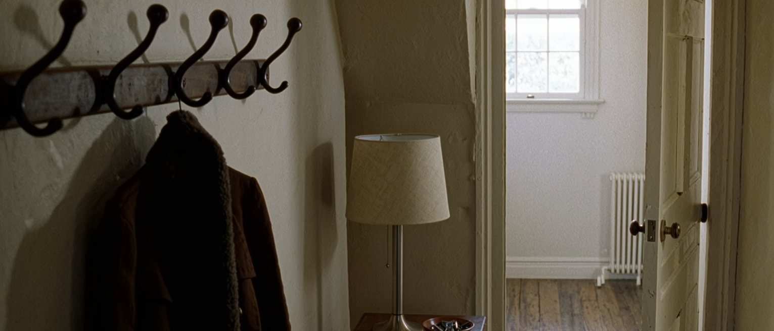 a hall seen close, a coat rack holding one coat and a row of empty hooks beside it, a hall table with a lamp and a bowl of keys beneath, the door on the latch