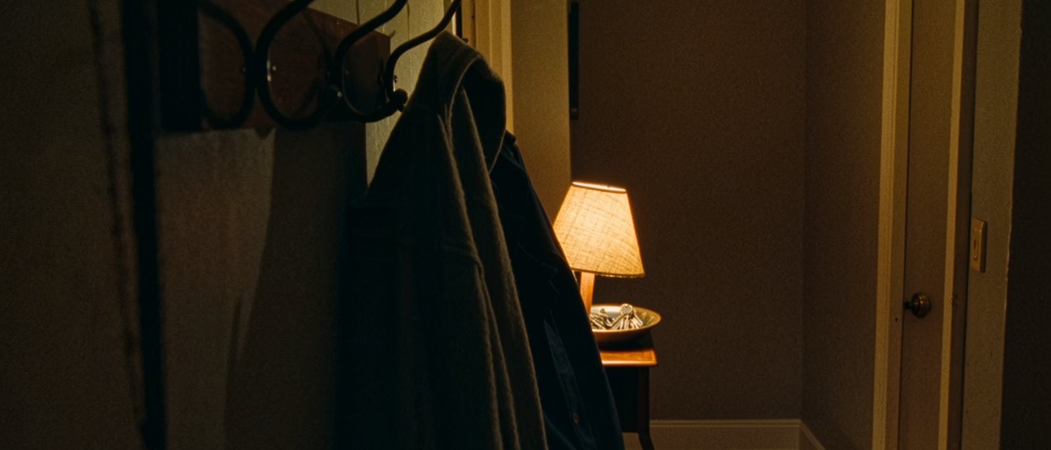 a hall seen close, a coat rack holding one coat and a row of empty hooks beside it, a hall table with a lamp and a bowl of keys beneath, the door on the latch