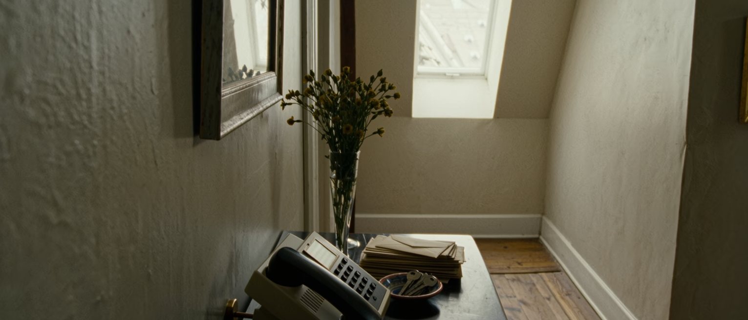 a hall table seen close with a telephone resting on its cradle beside a stack of unopened envelopes and a small dish of keys, a vase of stems going over, the door soft beyond