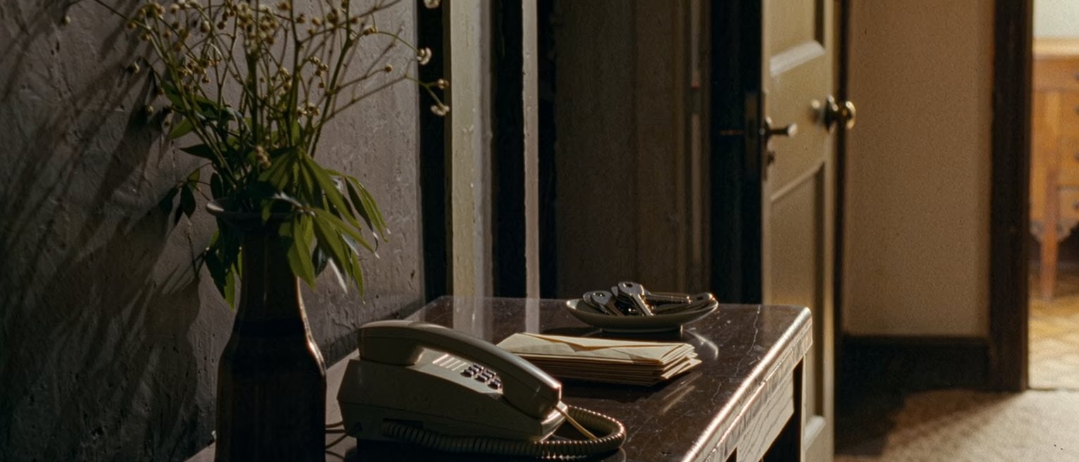 a hall table seen close with a telephone resting on its cradle beside a stack of unopened envelopes and a small dish of keys, a vase of stems going over, the door soft beyond