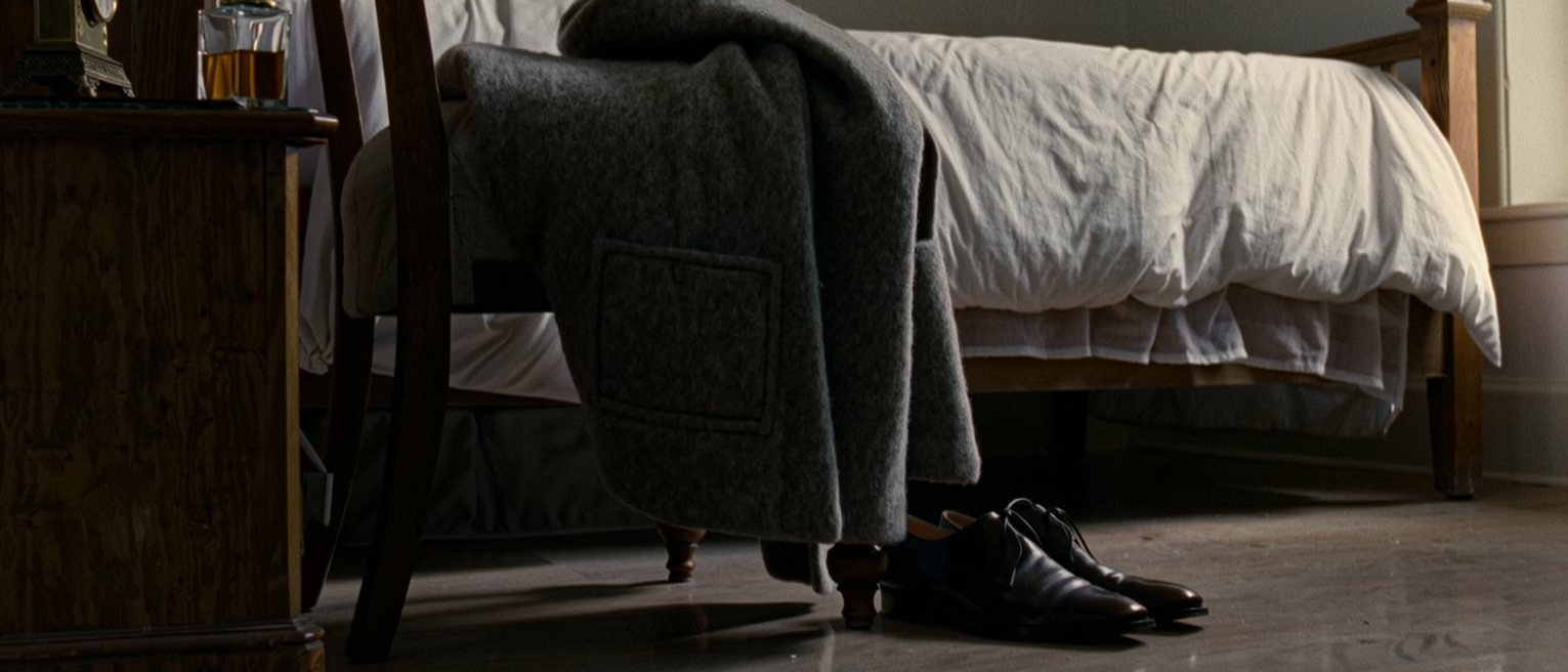 a bedroom seen close with a good coat laid over the back of the chair and a pair of shoes set out below it, a scent bottle and a small clock on the dresser, the bed made behind, the window soft beyond