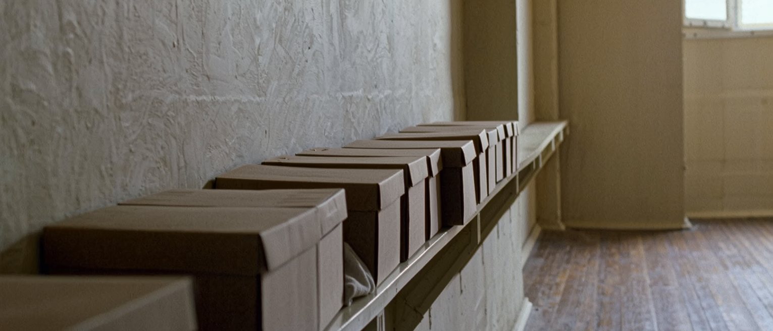 a long plain shelf along a quiet wall holding a row of identical unmarked boxes, one gap where a box has been lifted out, a folded cloth on the boards beneath