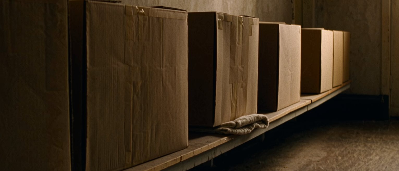 a long plain shelf along a quiet wall holding a row of identical unmarked boxes, one gap where a box has been lifted out, a folded cloth on the boards beneath