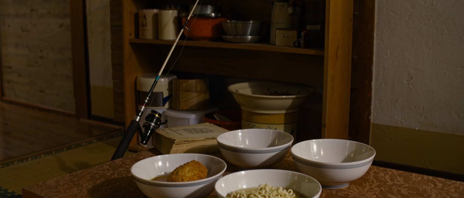 a low table seen close with instant noodles with a croquette on top in one bowl and four more bowls, a fishing rod leaning in the corner and an old pension book, the crowded shelves close behind