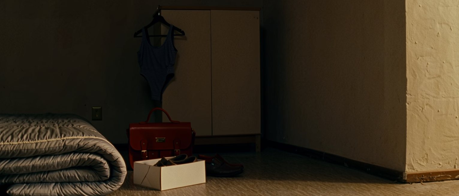 a small bedroom seen close with a futon rolled and a second one beside it, a pair of small shoes new in their box, a red satchel and a swimming costume drying on a hanger, the cupboard soft behind