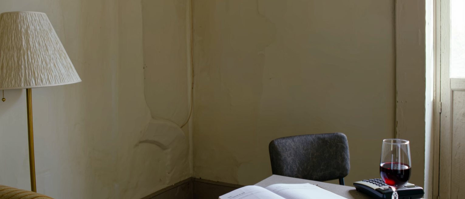 a hall table seen close with a telephone on it, a notebook open at a list of dates and a single glass of wine, the chair pulled in beside it and the lamp close behind