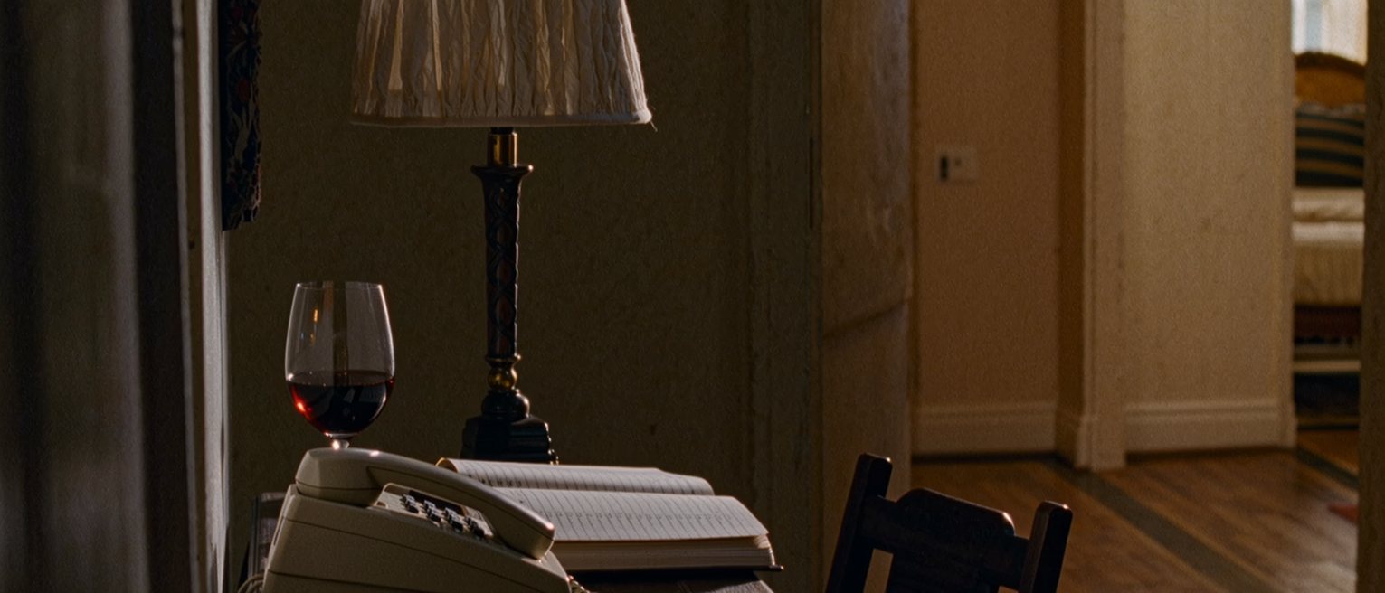 a hall table seen close with a telephone on it, a notebook open at a list of dates and a single glass of wine, the chair pulled in beside it and the lamp close behind